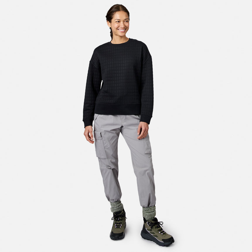 A woman in a black Free Country Women’s UltraFill Fleece Crew Neck Pullover Top, light gray cargo pants, gray socks, and green hiking boots smiles against a plain white background.