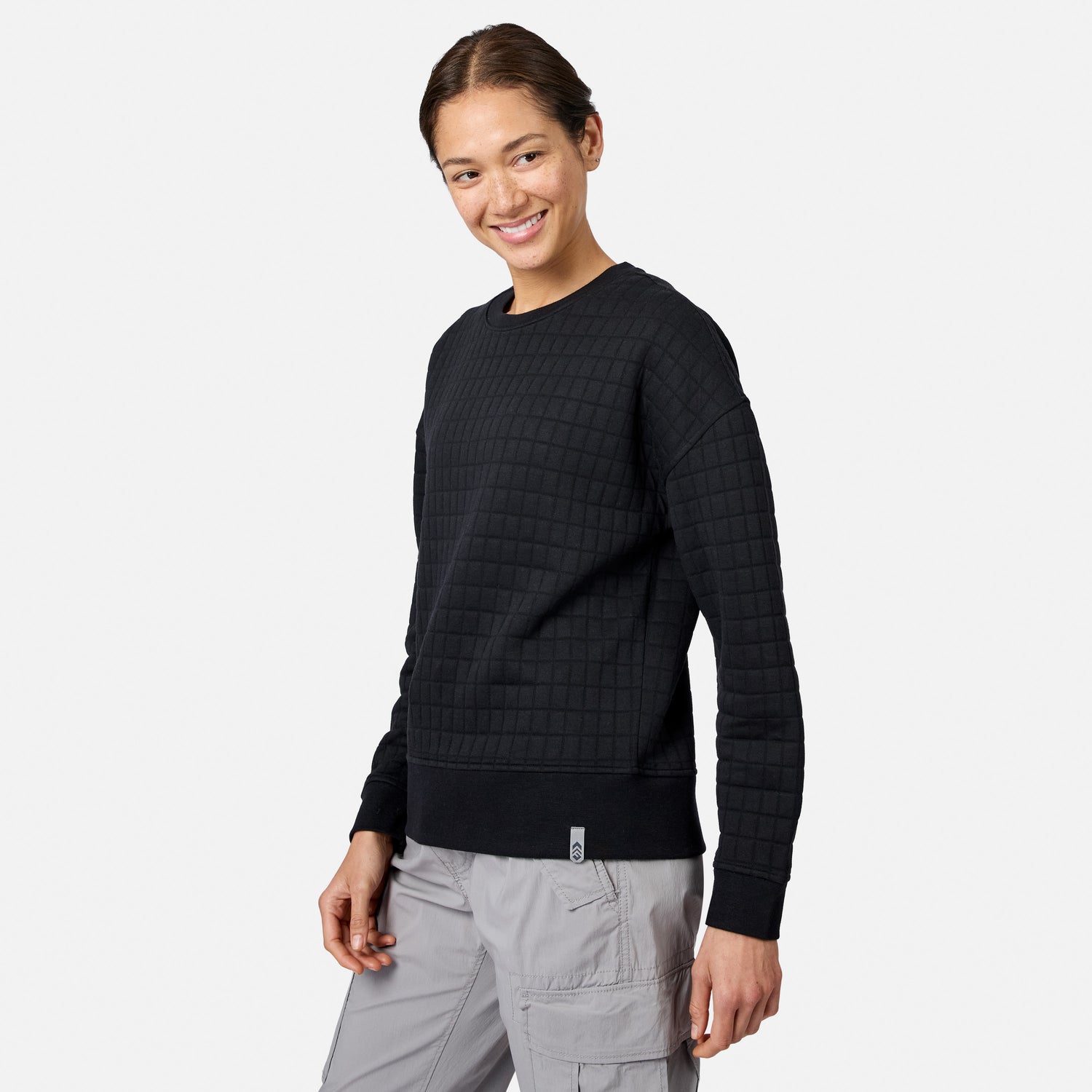 A person smiles in a black Women’s UltraFill Fleece Crew Neck Pullover Top by Free Country, paired with light gray cargo pants, standing against a plain white background.