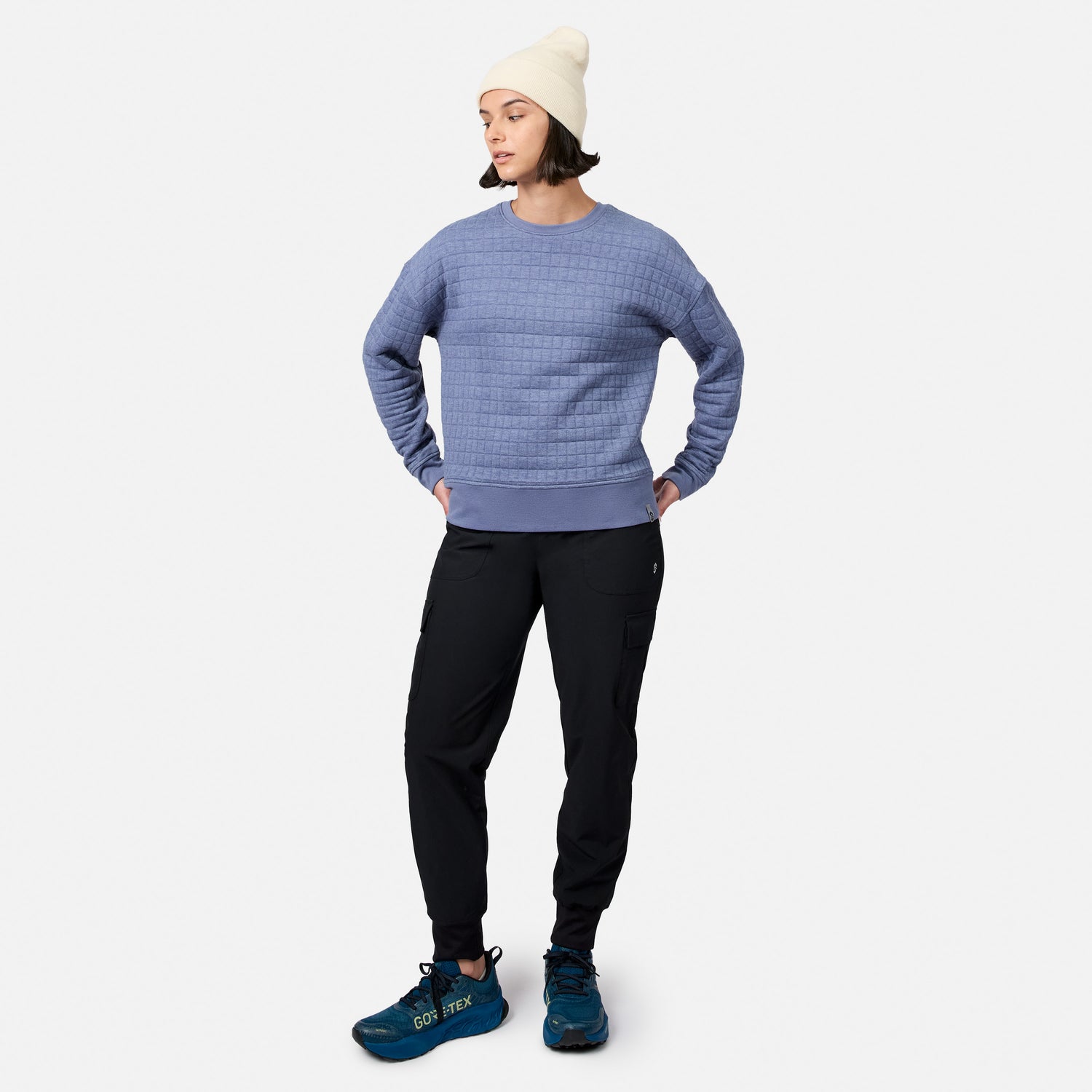 A person wears a cream beanie, Women’s UltraFill Fleece Crew Neck Pullover Top by Free Country, black cargo pants, and blue hiking shoes, standing hands behind back and looking left against a plain white background.