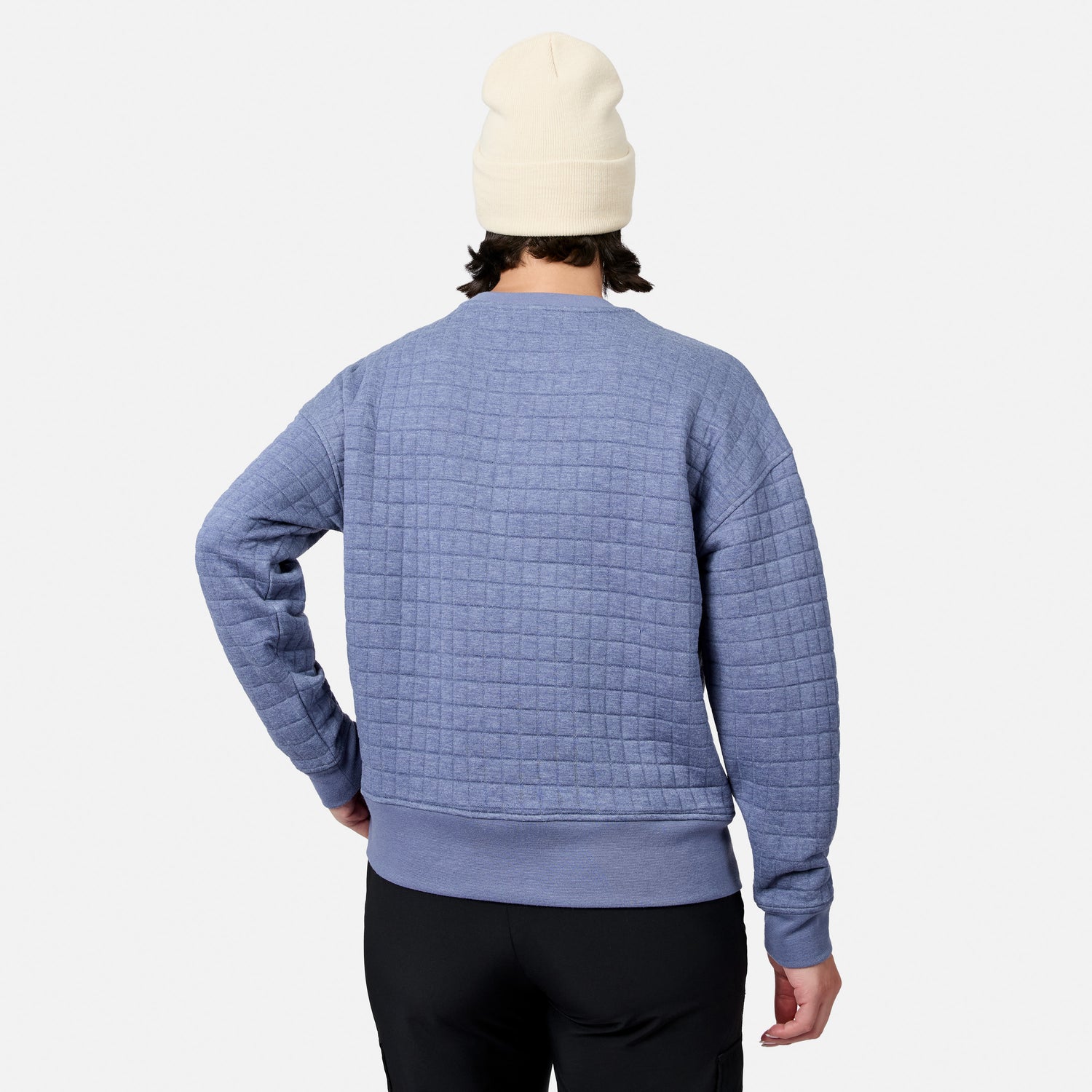 A person in a cream beanie, black pants, and a light blue Free Country Women’s UltraFill Fleece Crew Neck Pullover Top stands facing away from the camera against a white background.
