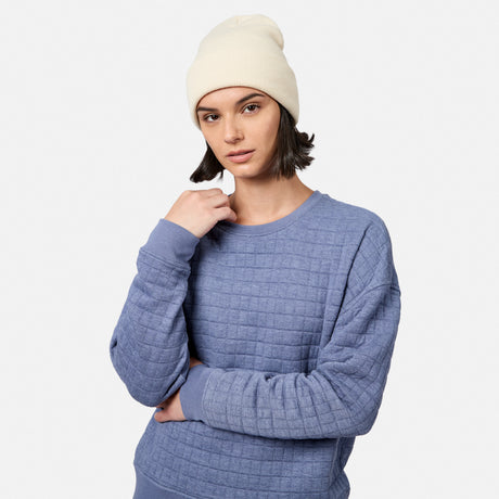 A person in a white beanie and an indigo Women’s UltraFill Fleece Crew Neck Pullover Top by Free Country stands against a plain white background, looking at the camera with a neutral expression.
