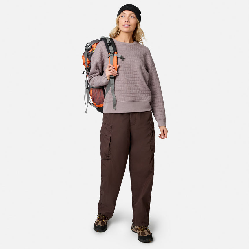 A smiling woman wearing a beanie, Free Country Women’s UltraFill Fleece Crew Neck Pullover Top in taupe, brown cargo pants, leopard print sneakers, and an orange hiking backpack poses against a plain white background.
