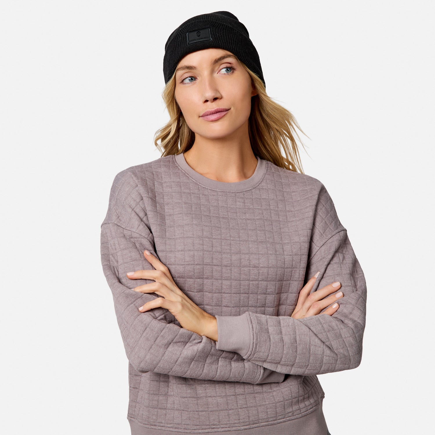 A woman with long blonde hair wears a black beanie and a mauve Free Country Women’s UltraFill Fleece Crew Neck Pullover Top, standing with arms crossed and looking aside, highlighting the top’s cozy comfort against a plain background.