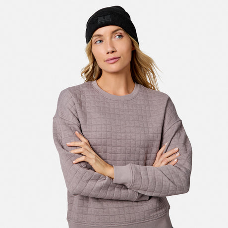 A woman with long blonde hair wears a black beanie and a mauve Free Country Women’s UltraFill Fleece Crew Neck Pullover Top, standing with arms crossed and looking aside, highlighting the top’s cozy comfort against a plain background.