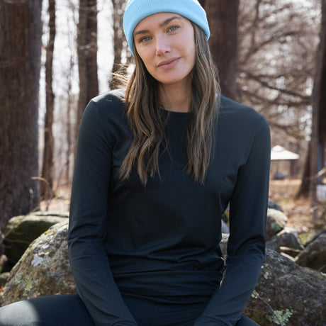 A person in a light blue beanie made from recycled materials and a black Free Country Women’s Microtech® Heat Cocarber® Coffee Base Layer Top sits on a rock outdoors, surrounded by trees with bare branches.
