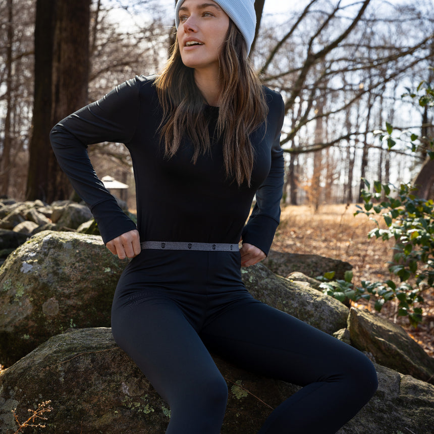 Wearing the Women’s Microtech® Heat Cocarber® Coffee Base Layer Bottom 2-Pack by Free Country, a woman sits on rocks outdoors among bare trees, smiling to the side in the sunshine while sporting a gray beanie and enjoying the comfort of her breathable black outfit.