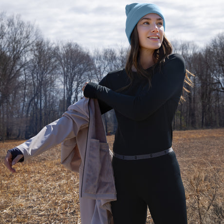 Wearing the Free Country Women’s Microtech® Heat Cocarber® Coffee Base Layer Top, a person in a blue beanie and black outfit stands outdoors on a field, smiling and holding a beige jacket. Dressed warmly in this innovative base layer, she enjoys the crisp air as leafless trees and a cloudy sky form the background.