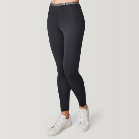 A person is shown from the waist down, wearing Free Country Women’s Microtech® Heat Cocarber® Coffee Base Layer Bottoms in black, paired with white sneakers, standing against a plain white background.