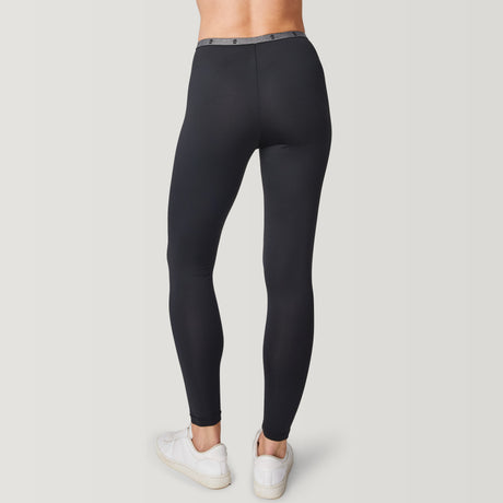 A person is shown from the back against a plain light background, wearing black Women’s Microtech® Heat Cocarber® Coffee Base Layer Bottom leggings by Free Country paired with white sneakers. The high-waisted leggings have a fitted silhouette and are made from breathable, four-way stretch fabric for maximum comfort and flexibility. The person’s upper body is not visible.