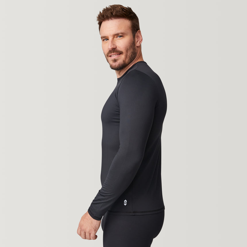 A man with short brown hair and a beard wears the Free Country Men’s Microtech® Heat Cocarber® Coffee Base Layer Top, a fitted black long-sleeve shirt made from recycled materials, along with black pants, standing sideways and looking at the camera against a plain light background.