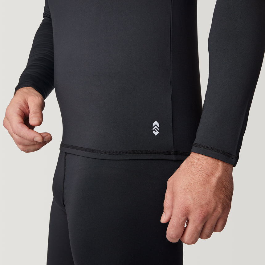 A person wearing the Free Country Men’s Microtech® Heat Cocarber® Coffee Base Layer Top, a fitted black long-sleeve shirt made with recycled materials and featuring a small white logo near the hem; only their torso and hands are visible.