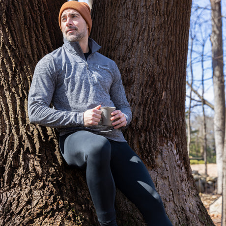A man in a gray jacket, Free Country Men’s Microtech® Heat Cocarber® Coffee Base Layer Bottoms, and a brown beanie leans against a large tree while holding a mug. It appears to be a sunny day with bare trees in the background.