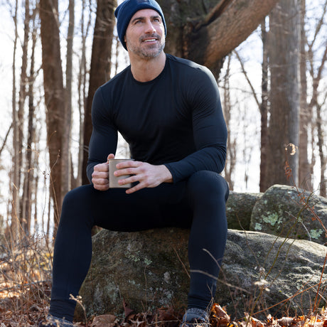 Wearing the Free Country Men’s Microtech® Heat Cocarber® Coffee Base Layer Top, a man in black athletic apparel and a blue beanie sits on forest rocks, holding a mug and smiling as bare trees and sunlight fill the background.