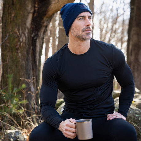 A man in a navy beanie and the Free Country Men’s Microtech® Heat Cocarber® Coffee Base Layer Top sits outdoors holding a metal mug, with trees and sunlight in the background.