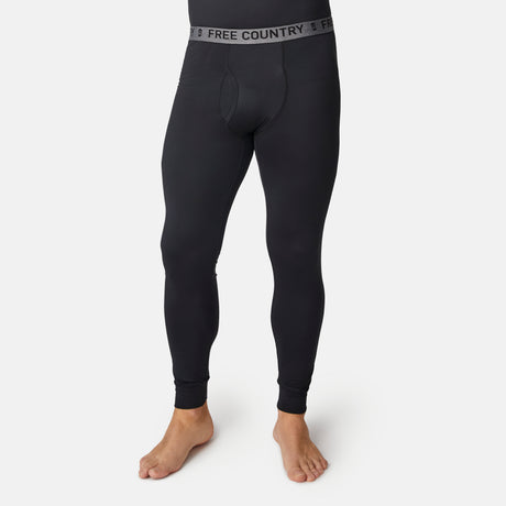 A person stands barefoot against a white background wearing Free Country Men’s Microtech® Heat Cocarber® Coffee Base Layer Bottoms in black, featuring a gray waistband with the brand’s logo, moisture-wicking technology, and four-way stretch.