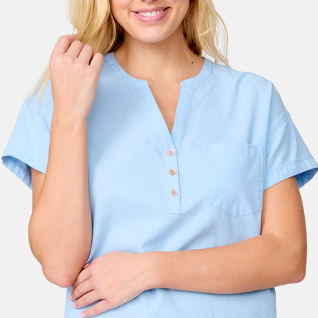 A woman with loose blonde hair smiles, one hand raised, wearing the Free Country Women's Easy Breezy Shirt in light blue cotton-linen with short sleeves, front pocket, and buttoned neckline. She stands against a plain light gray background.