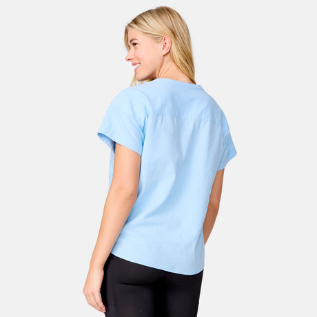 A woman with long blonde hair models the Free Country Women's Easy Breezy Shirt, a pastel striped V-neck short sleeve top, paired with black pants. She stands against a plain light background, smiling over her shoulder.