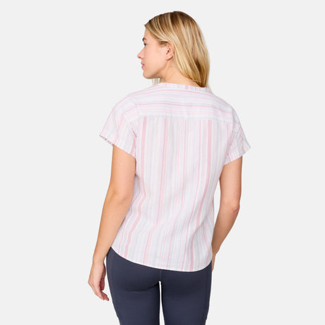 A woman with blonde hair wears the Free Country Women's Easy Breezy Shirt, featuring pastel stripes and a V-neck with short sleeves, paired with dark pants. She stands with her back to the camera, glancing over her left shoulder against a white background.
