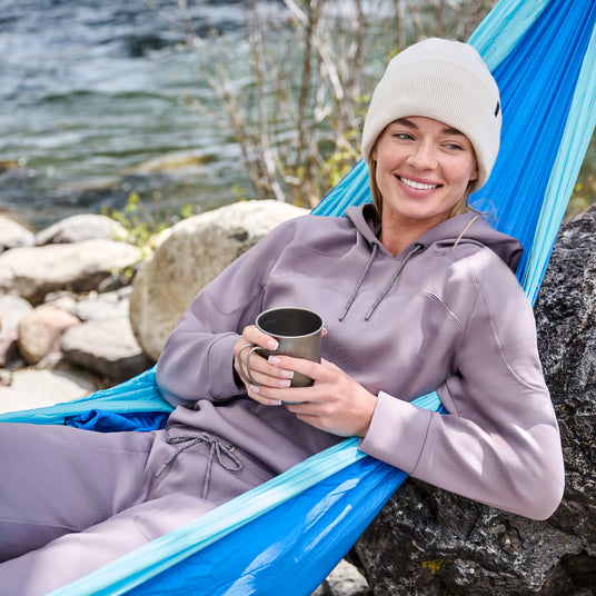 Wearing the Free Country Women’s Sueded Lush Jogger, a person in a light purple hoodie and beige beanie relaxes in a blue hammock by a river, smiling with a metal cup amongst rocks and trees.