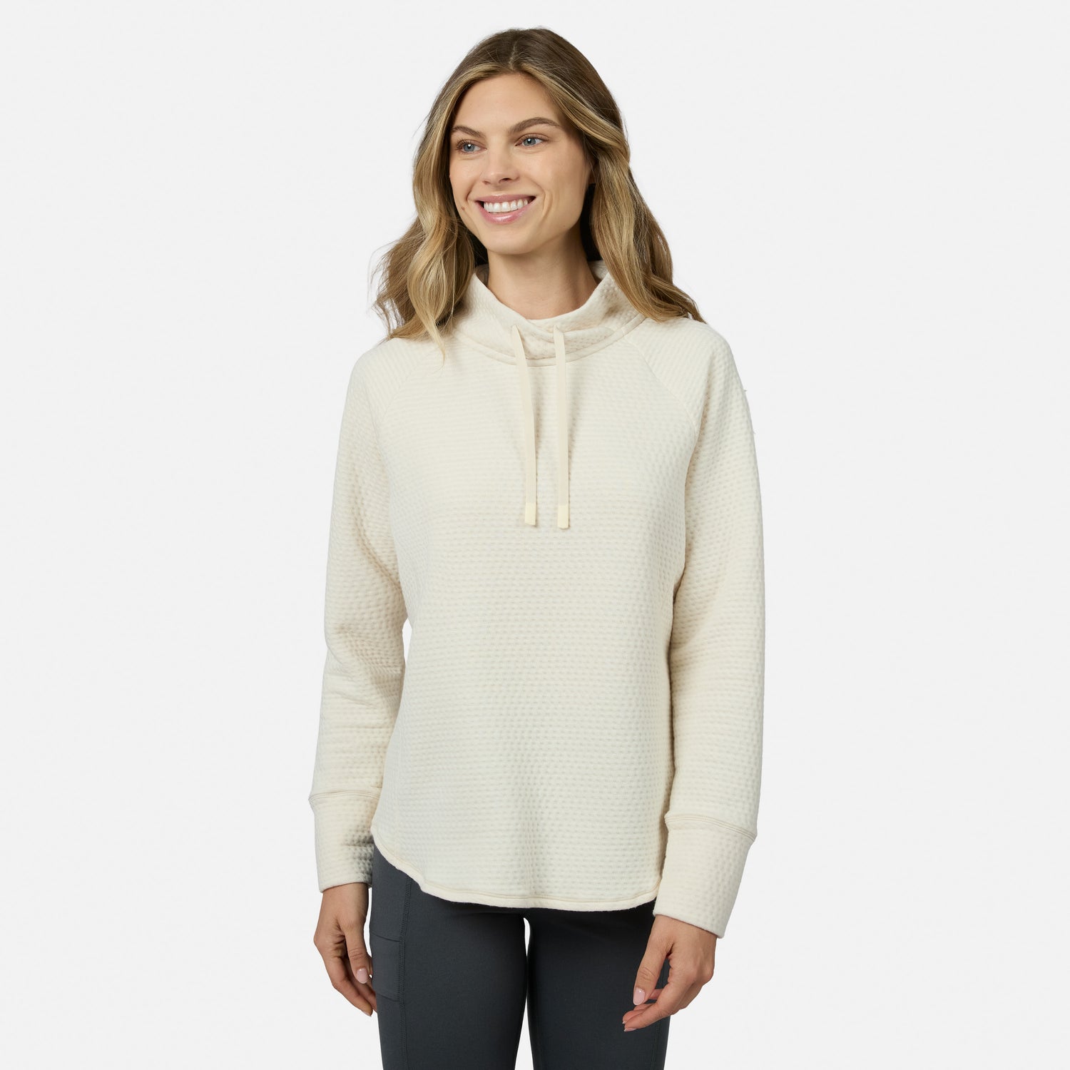 A smiling woman wears a cream-colored Free Country Women’s UltraFill Cowl Neck Top featuring a textured, seamless quilting pattern and a drawstring.