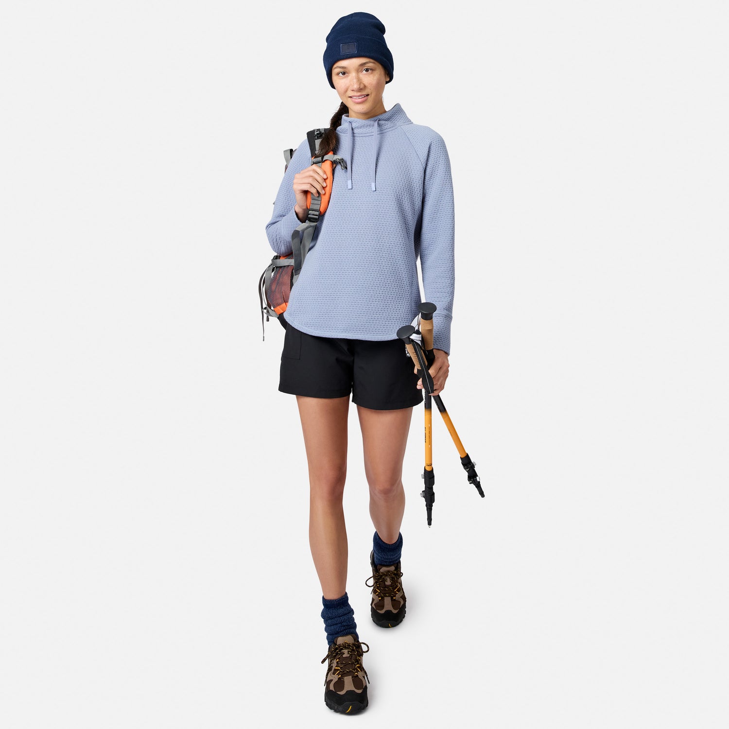 A woman wearing a blue Free Country Women’s UltraFill Cowl Neck Top, shorts, and a beanie, carrying a backpack and hiking poles.