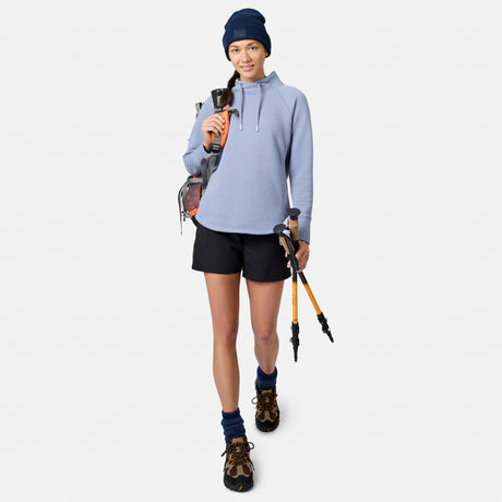 A woman wearing a blue Free Country Women’s UltraFill Cowl Neck Top, shorts, and a beanie, carrying a backpack and hiking poles.