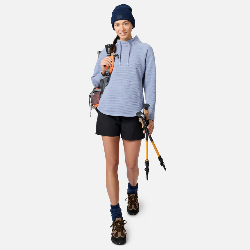 A woman wearing a blue Free Country Women’s UltraFill Cowl Neck Top, shorts, and a beanie, carrying a backpack and hiking poles.