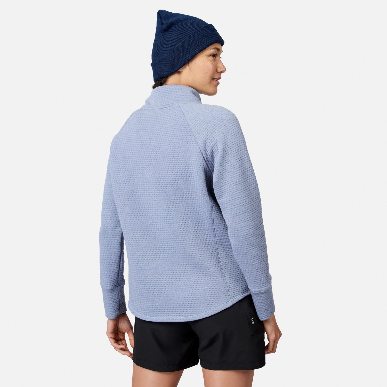 A person wearing a blue textured Free Country Women’s UltraFill Cowl Neck Top, black shorts, and a navy blue beanie.
