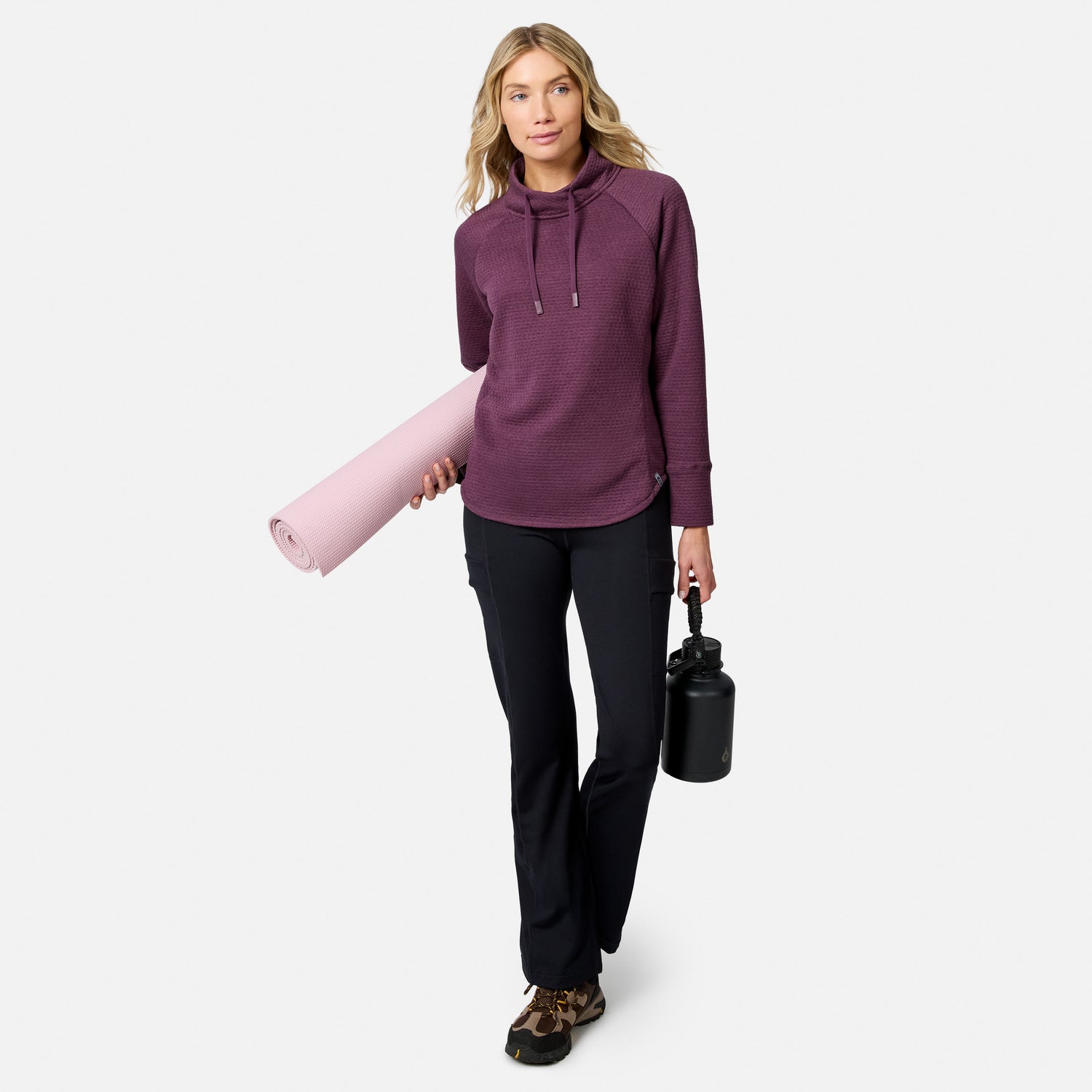 A woman wearing a purple Free Country Women’s UltraFill Cowl Neck Top and black pants carries a light pink yoga mat and a black water bottle.