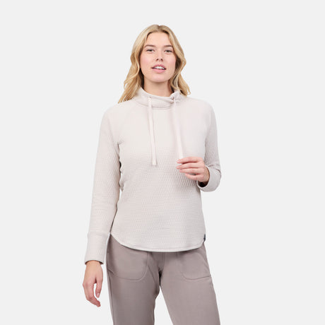 A woman with long blonde hair wears the Free Country Women's UltraFill Cowl Neck Top with seamless quilting and light polyester fill, paired with taupe pants, standing against a plain white background facing forward.