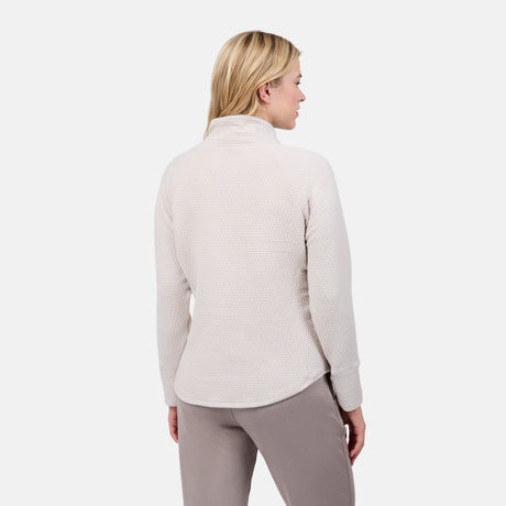 A blonde woman is seen from the back wearing the Free Country Women's UltraFill Cowl Neck Top with light polyester fill and taupe pants, highlighting seamless quilting against a plain white background.
