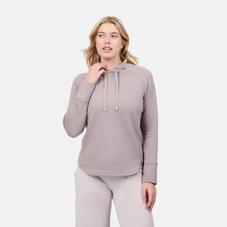 A woman models the Free Country Women's UltraFill Cowl Neck Top, featuring a high collar with drawstrings and seamless quilting, as she stands against a white background with one hand near her neck.