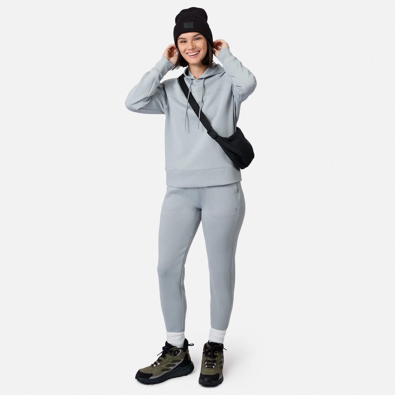 Smiling woman in a Free Country Women’s Sueded Lush Jogger and matching hoodie, black beanie, crossbody bag, white socks, and hiking boots stands with hands at her hoodie against a plain white background.