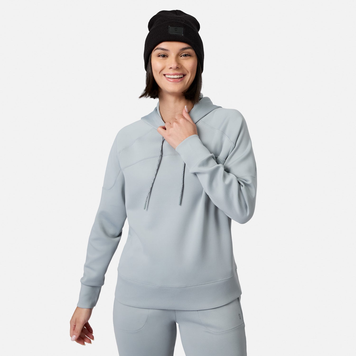 A woman in a Free Country Women’s Sueded Lush Pullover Hoodie and matching pants smiles at the camera, wearing a black beanie and adjusting her hood. The cozy hoodie pops against the plain white background.