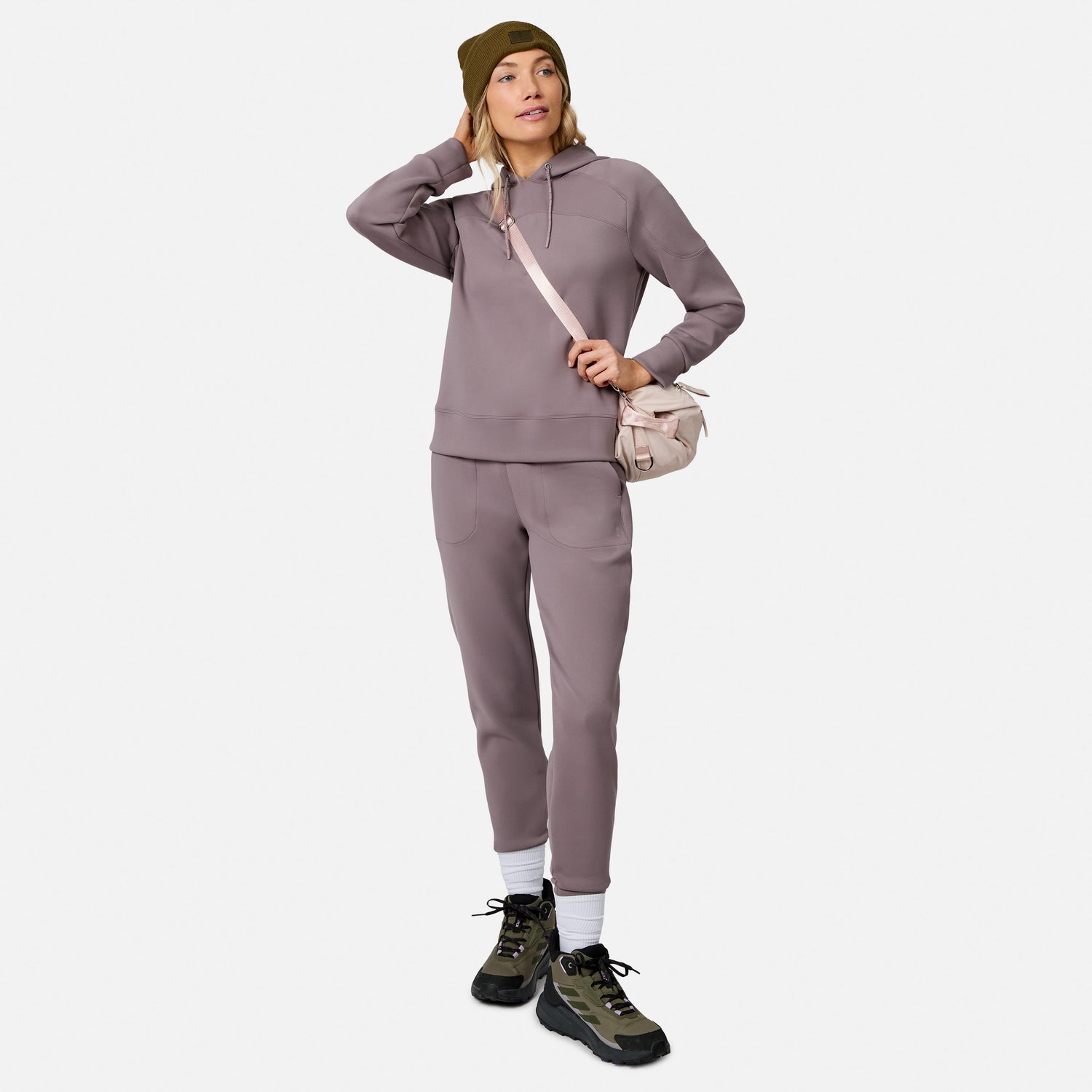 Woman wearing the Free Country Women’s Sueded Lush Pullover Hoodie, mauve joggers, olive beanie, hiking boots, white socks, and a light pink crossbody bag, standing against a plain white background.