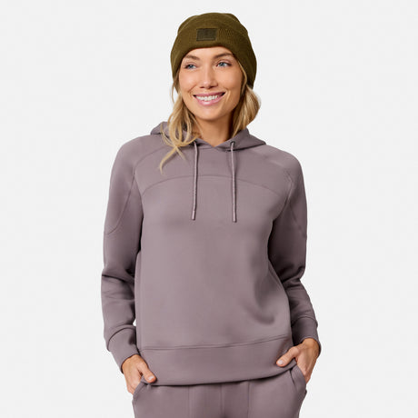 Smiling woman wears the Free Country Women’s Sueded Lush Pullover Hoodie in taupe with matching pants and an olive green beanie, hands in pockets, against a plain white background.