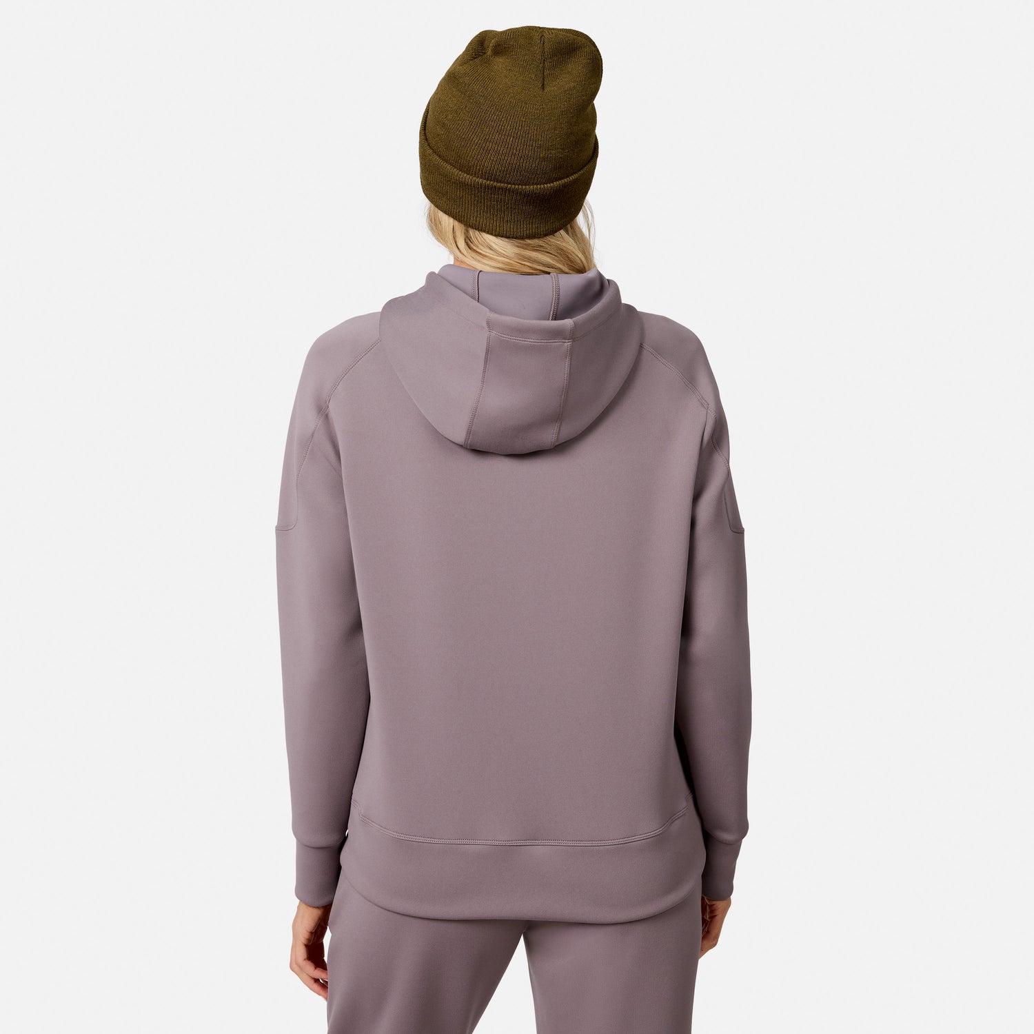 A woman is seen from the back wearing a mauve Free Country Women’s Sueded Lush Pullover Hoodie with matching pants and a brown beanie, standing against a plain white background.