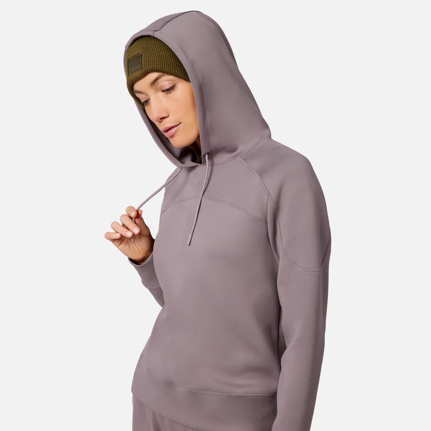 A woman wears the Free Country Women’s Sueded Lush Pullover Hoodie with the hood up over a green beanie, looking down and holding the drawstring. The cozy sweatshirt stands out against a plain white background.