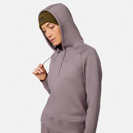 A woman wears the Free Country Women’s Sueded Lush Pullover Hoodie with the hood up over a green beanie, looking down and holding the drawstring. The cozy sweatshirt stands out against a plain white background.
