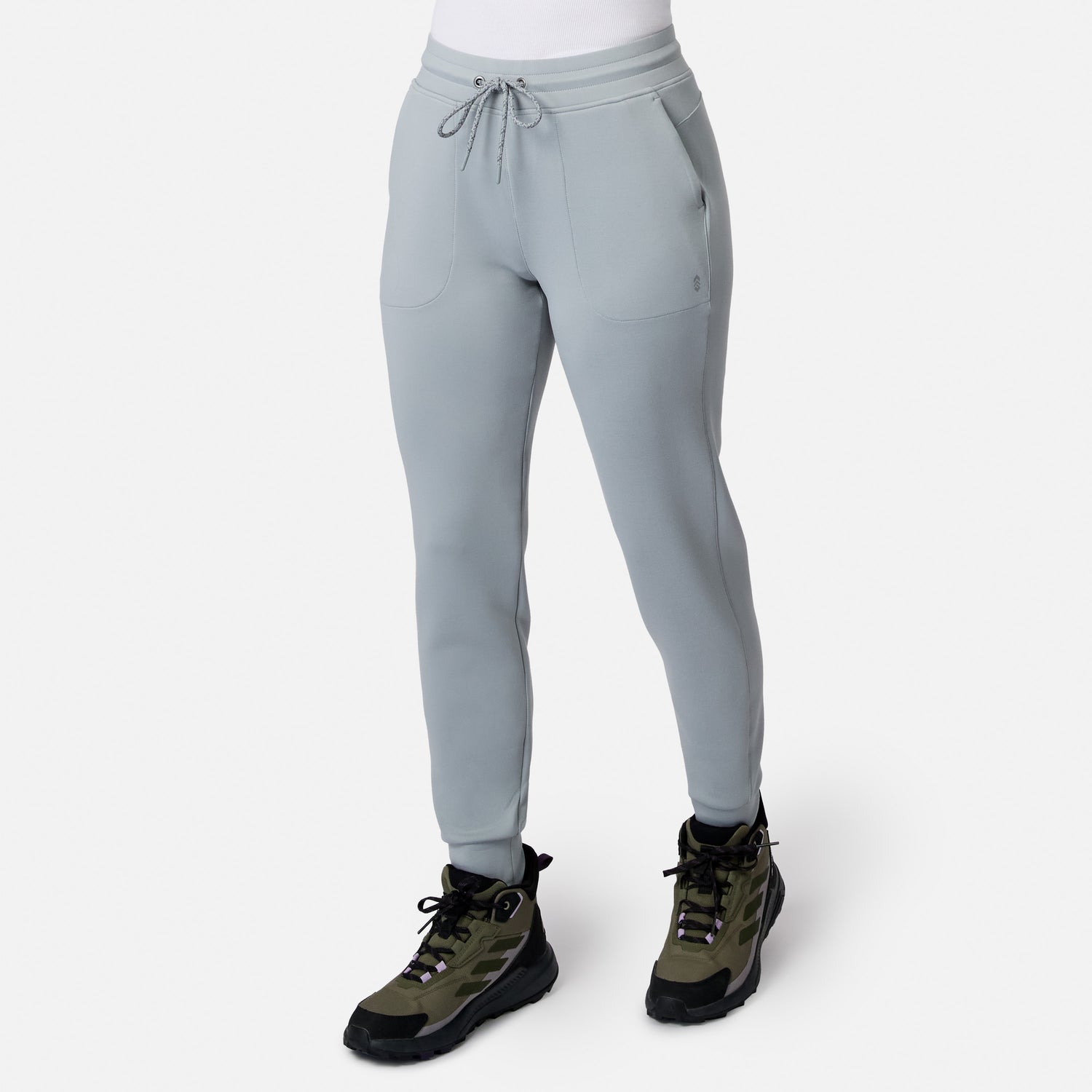 A woman wearing Free Country Women’s Sueded Lush Jogger in Lichen with olive green hiking boots stands against a plain white background.