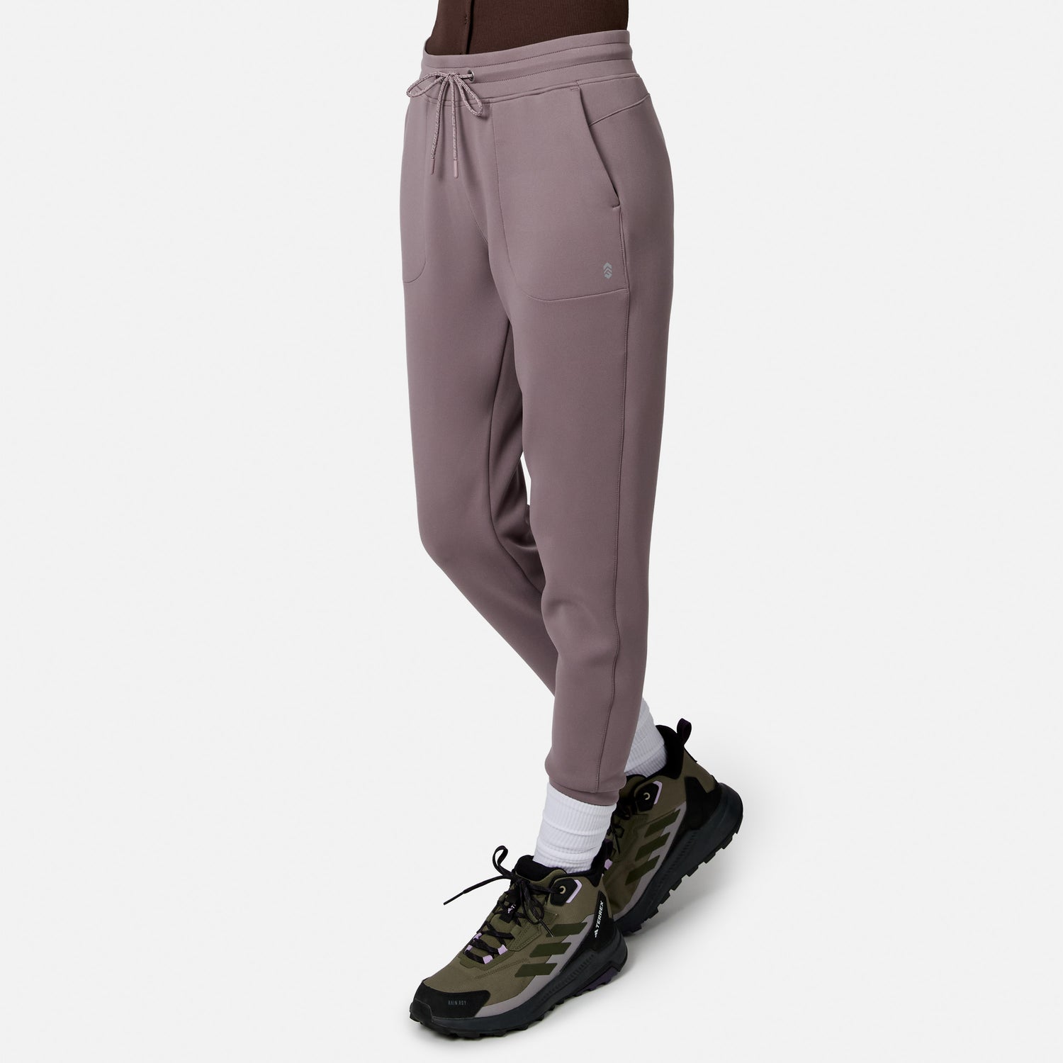 Person wearing Free Country Women’s Sueded Lush Jogger in mauve with drawstring waist and pockets, white socks, and green-black athletic shoes, standing against a plain white background. Pants offer comfort and flexibility with 4-way stretch.
