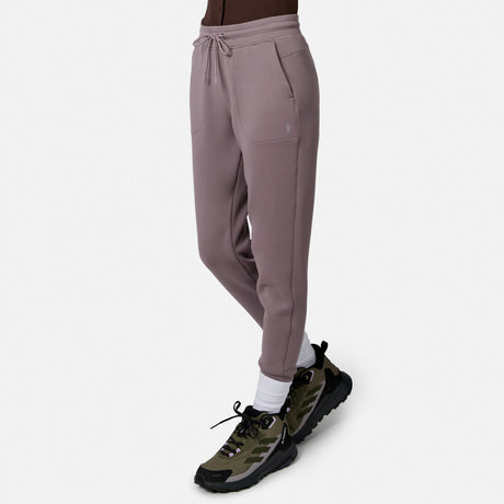Person wearing Free Country Women’s Sueded Lush Jogger in mauve with drawstring waist and pockets, white socks, and green-black athletic shoes, standing against a plain white background. Pants offer comfort and flexibility with 4-way stretch.