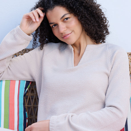 A woman with curly hair, wearing the Free Country Women's Ripple Knit Long Sleeve Pullover Top, sits on a wicker bench, resting her arm on a colorful striped pillow and smiling softly at the camera.