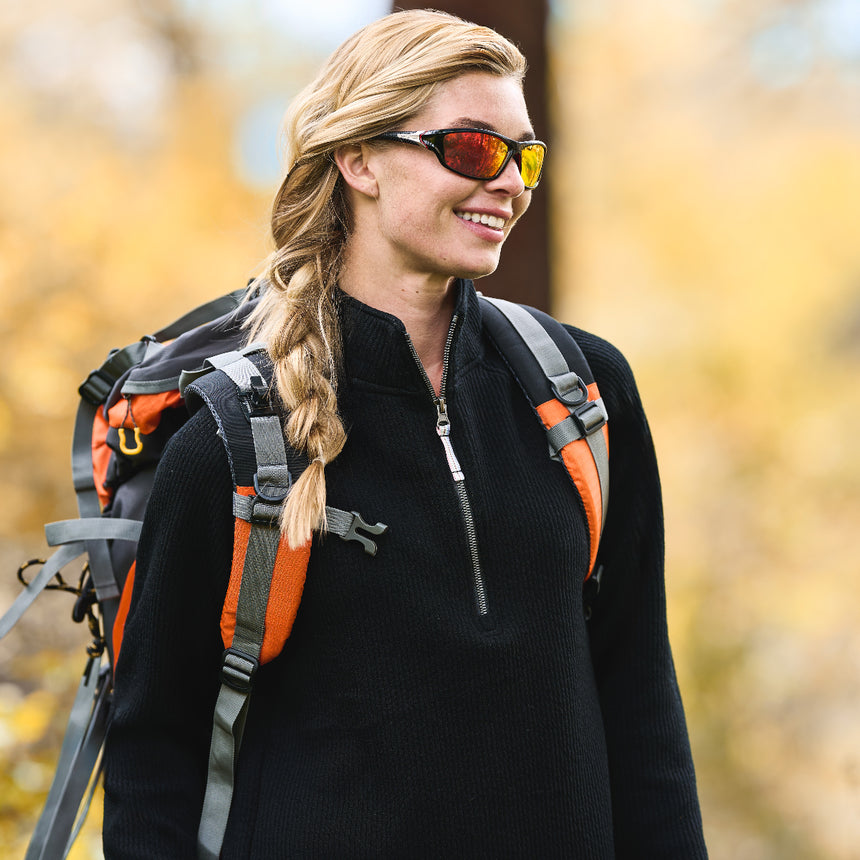 A woman with a long blonde braid, in sunglasses and a Free Country Women’s Ripple Knit Half Zip Top, smiles outdoors carrying an orange and gray backpack against a blurred yellow background.