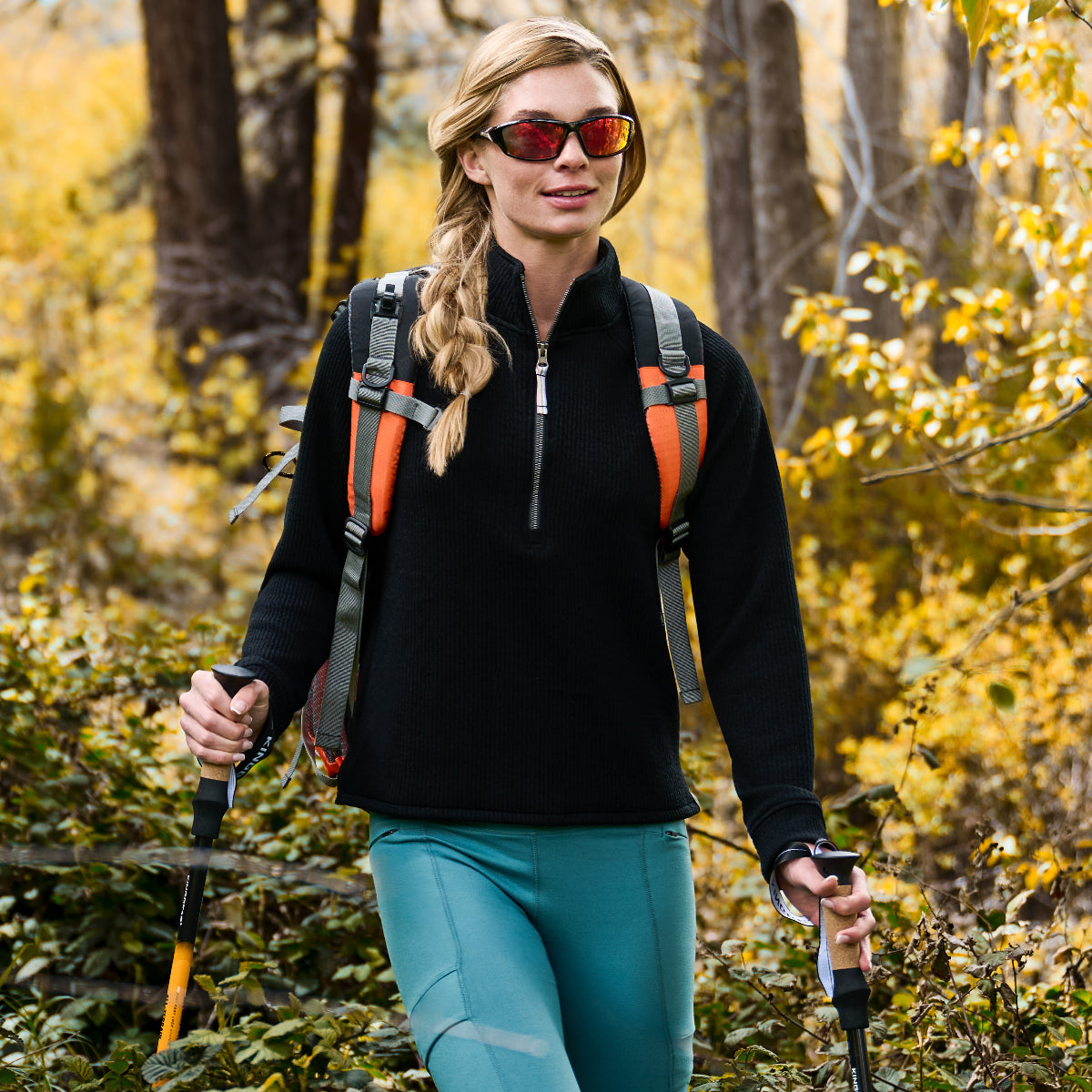 A woman hikes in the forest wearing the Free Country Women’s Ripple Knit Half Zip Top and teal pants, layered for style. She pairs red sunglasses, a backpack with orange straps, and trekking poles among green foliage and yellow autumn trees.