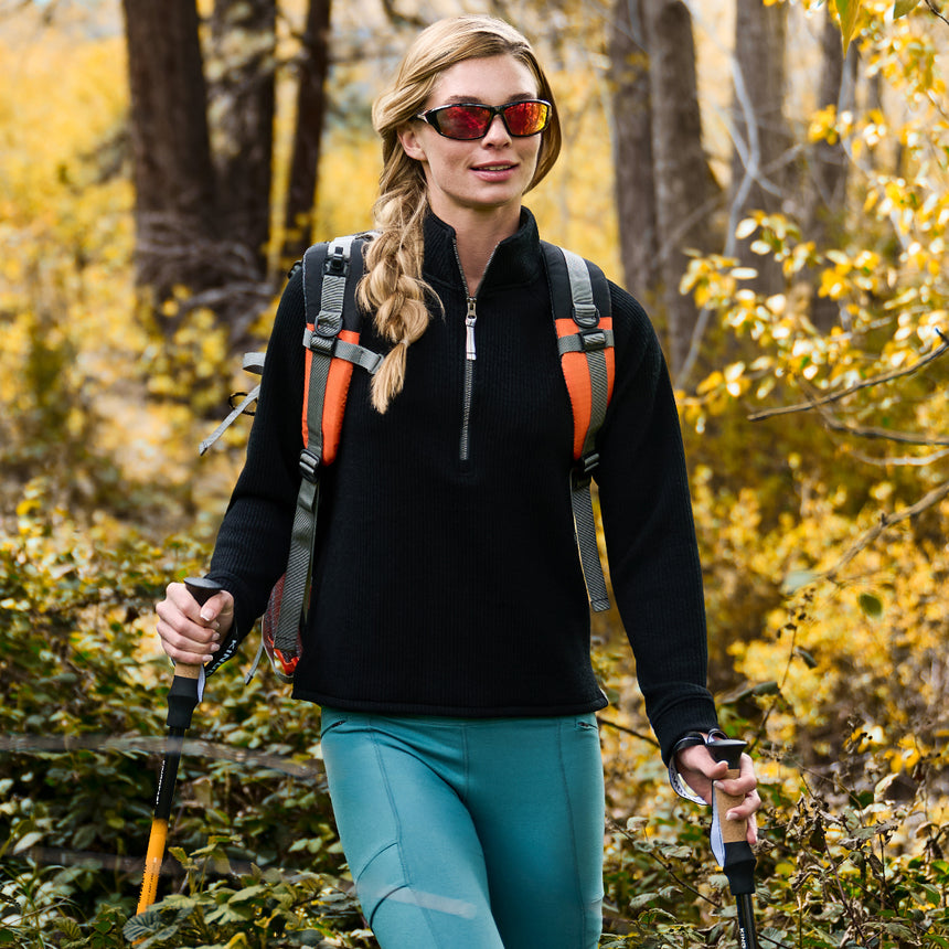 A woman hikes in the forest wearing the Free Country Women’s Ripple Knit Half Zip Top and teal pants, layered for style. She pairs red sunglasses, a backpack with orange straps, and trekking poles among green foliage and yellow autumn trees.
