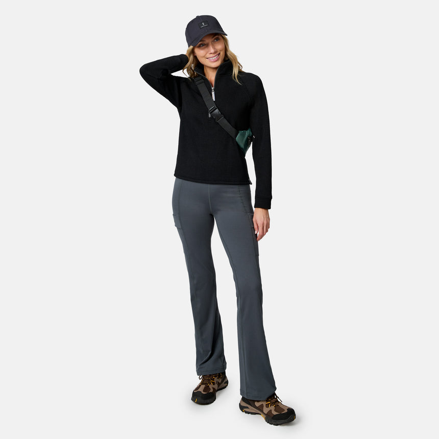 A woman in a Free Country Women’s Ripple Knit Half Zip Top (black), gray pants, brown hiking shoes, black crossbody bag, and black cap stands smiling with one hand on her head against a plain light background.