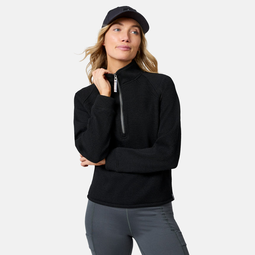 A woman models the Free Country Women’s Ripple Knit Half Zip Top in black, paired with gray pants and a black cap, standing against a plain background—ideal for casual layering.