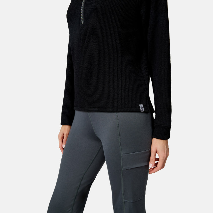 A woman wears the Free Country Women’s Ripple Knit Half Zip Top in black, paired with gray leggings with side pockets. Only her midsection and upper legs are shown against a white background, highlighting casual, layered style.