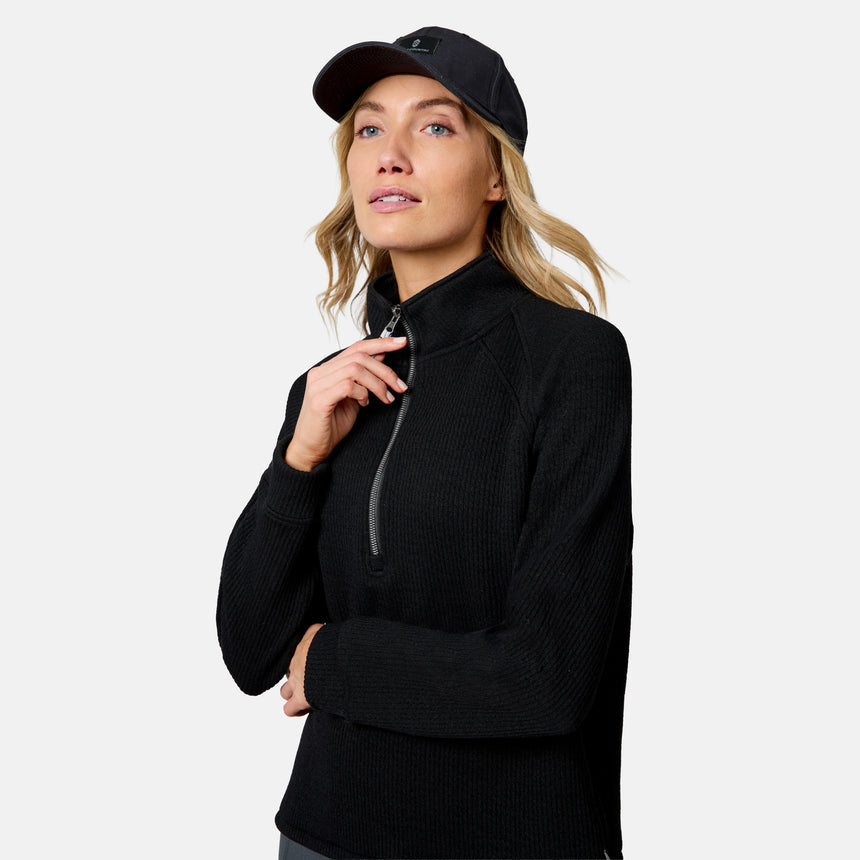 A woman wears the Free Country Women’s Ripple Knit Half Zip Top and a black baseball cap, looking upward with a neutral expression against a plain light background, highlighting her casual layered style.